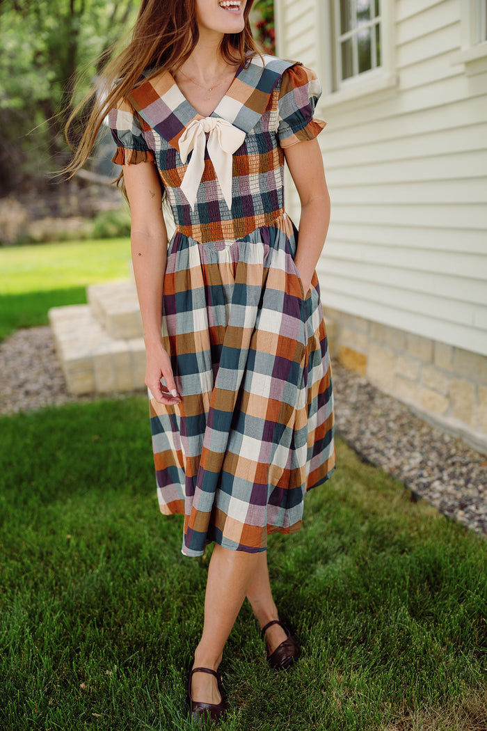 A woman in the Alice Dress in Maize Gingham by Ivy City Co, featuring puff sleeves and a smocked bodice, stands on grass near a white house. She is smiling with her long hair down and has her hands in her pockets.