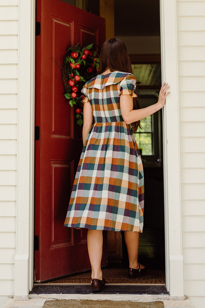 A woman wearing the Ivy City Co Alice Dress in Maize Gingham, featuring puff sleeves and a smocked bodice, enters a house through a red door adorned with a wreath, her back to the camera.