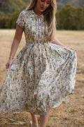 A woman stands outdoors on dry grass, holding one side of her skirt as she wears the Ivy City Co Amber Dress in Autumn Bouquet—a flowing, short-sleeved floral tea-length style that is both elegant and nursing friendly—with trees and a blurred background behind her.