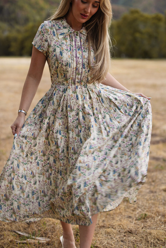 A woman stands outdoors on dry grass, holding one side of her skirt as she wears the Ivy City Co Amber Dress in Autumn Bouquet—a flowing, short-sleeved floral tea-length style that is both elegant and nursing friendly—with trees and a blurred background behind her.