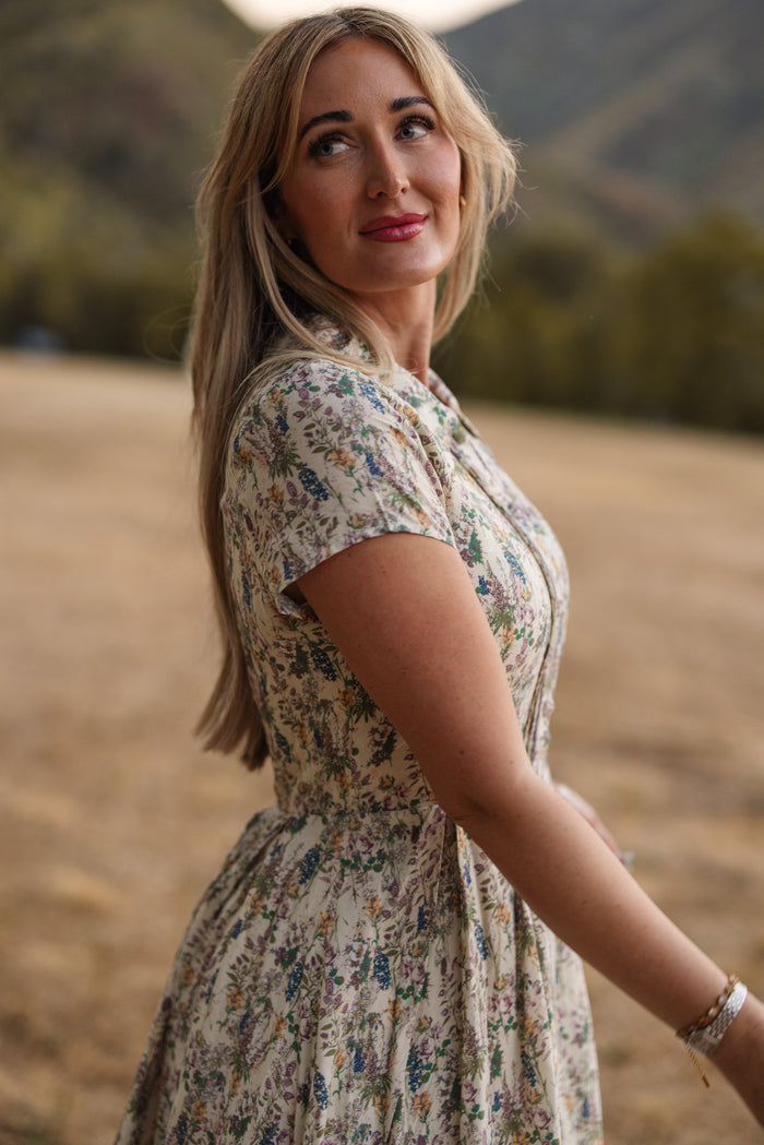 A woman with long blonde hair stands outdoors in the Amber Dress in Autumn Bouquet by Ivy City Co—a tea-length floral dress that is both nursing friendly and perfect for a stroll on a sunny day. The background features blurred grass and green hills as she gazes to the side.