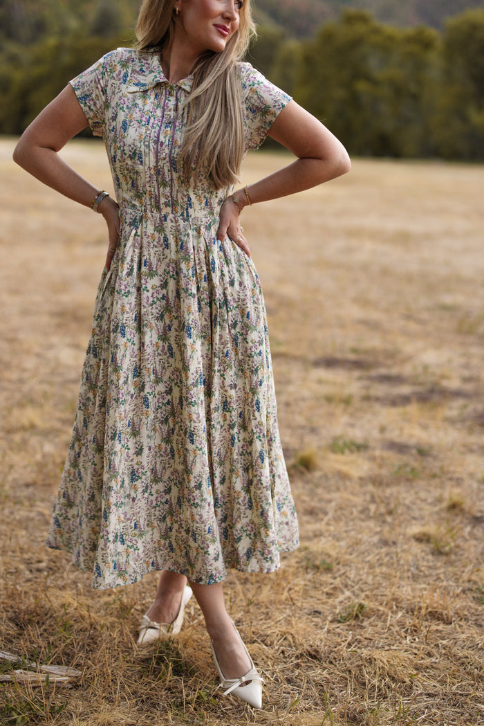 A person stands in a dry grassy field with trees in the background, hands resting on their hips and wearing light-colored heels, dressed in the Ivy City Co Amber Dress in Autumn Bouquet—a floral, short-sleeve, button-up tea-length dress that is nursing friendly and combines style with comfort.