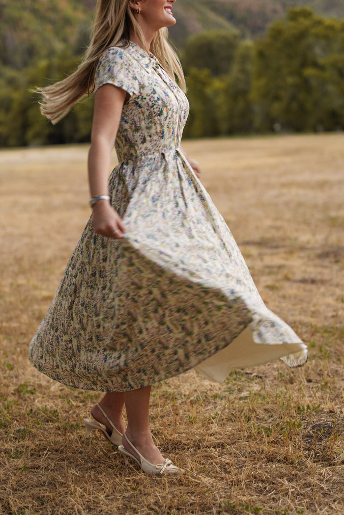 Wearing the Ivy City Co Amber Dress in Autumn Bouquet—a nursing friendly, floral tea-length design—a woman twirls on dry grass in an open field, with softly blurred trees and hills in the background.