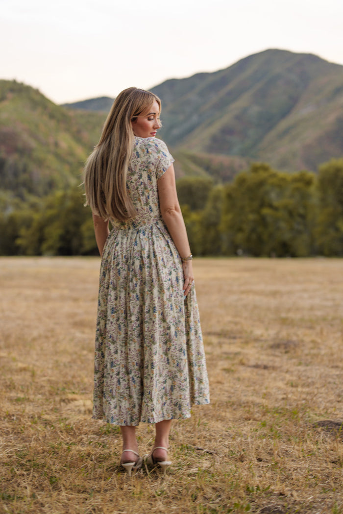 A woman with long blonde hair, wearing the Ivy City Co Amber Dress in Autumn Bouquet—a tea-length floral dress—stands in a dry grassy field, facing away toward green mountains and trees under a soft sky.