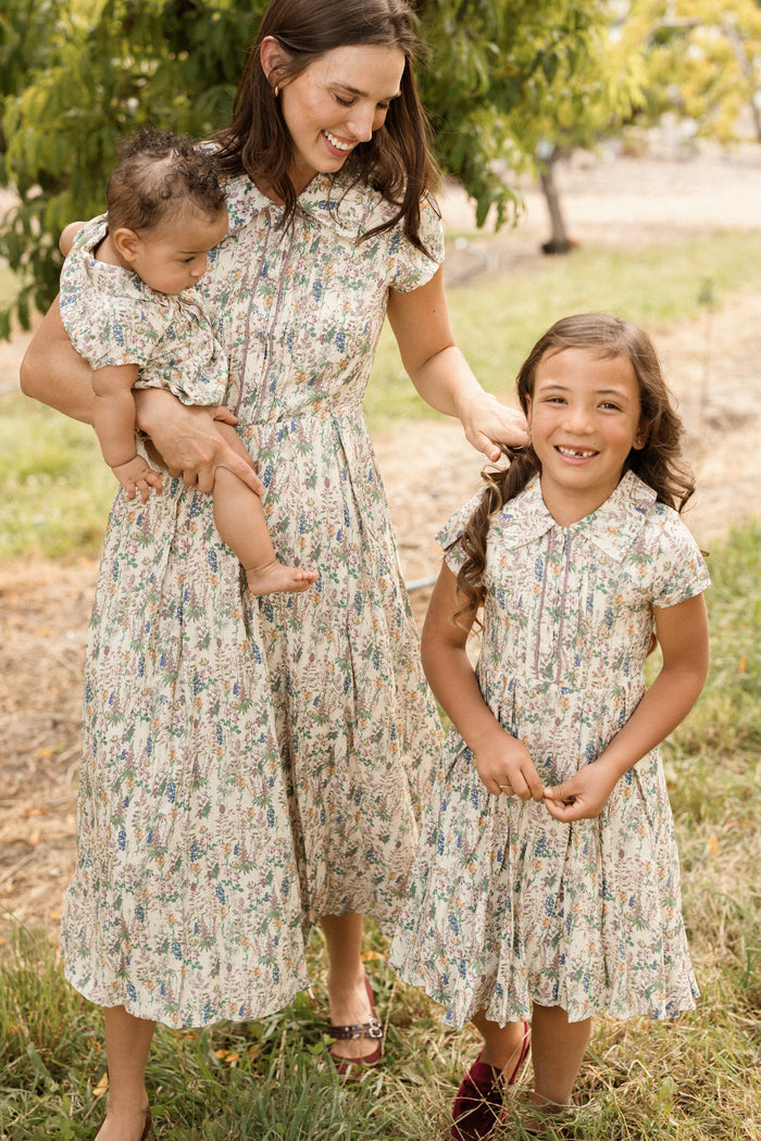 A woman holds a baby and stands beside a young girl, all three smiling in matching Ivy City Co Amber Dresses in Autumn Bouquet. Their floral tea-length outfits stand out against the sunlit, grassy backdrop with trees, while the woman's nursing-friendly dress adds both style and practicality for mothers.