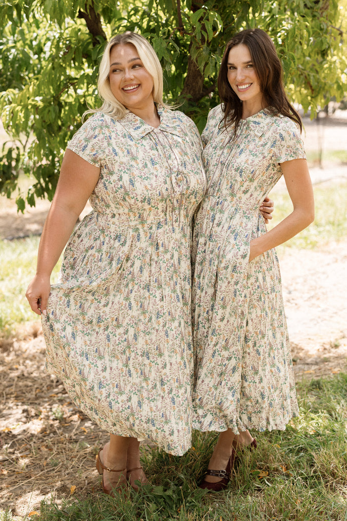 Two women stand side by side outdoors, smiling and wearing matching Ivy City Co Amber Dresses in Autumn Bouquet with short sleeves. They hold the skirts of their tea-length dresses amidst green trees and sunlight in the background.