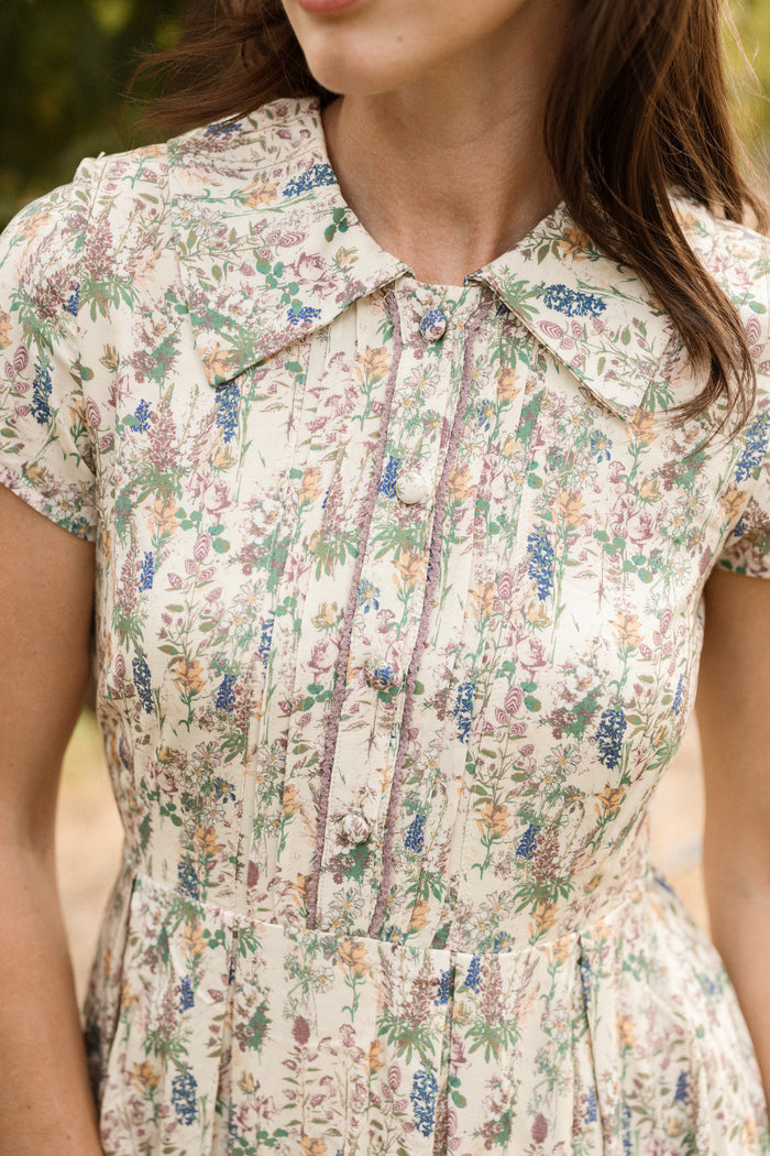 A woman wearing the Ivy City Co Amber Dress in Autumn Bouquet—a vintage-inspired, nursing-friendly tea-length dress with a floral print and pointed collar—stands outdoors. Her face is partially out of frame.