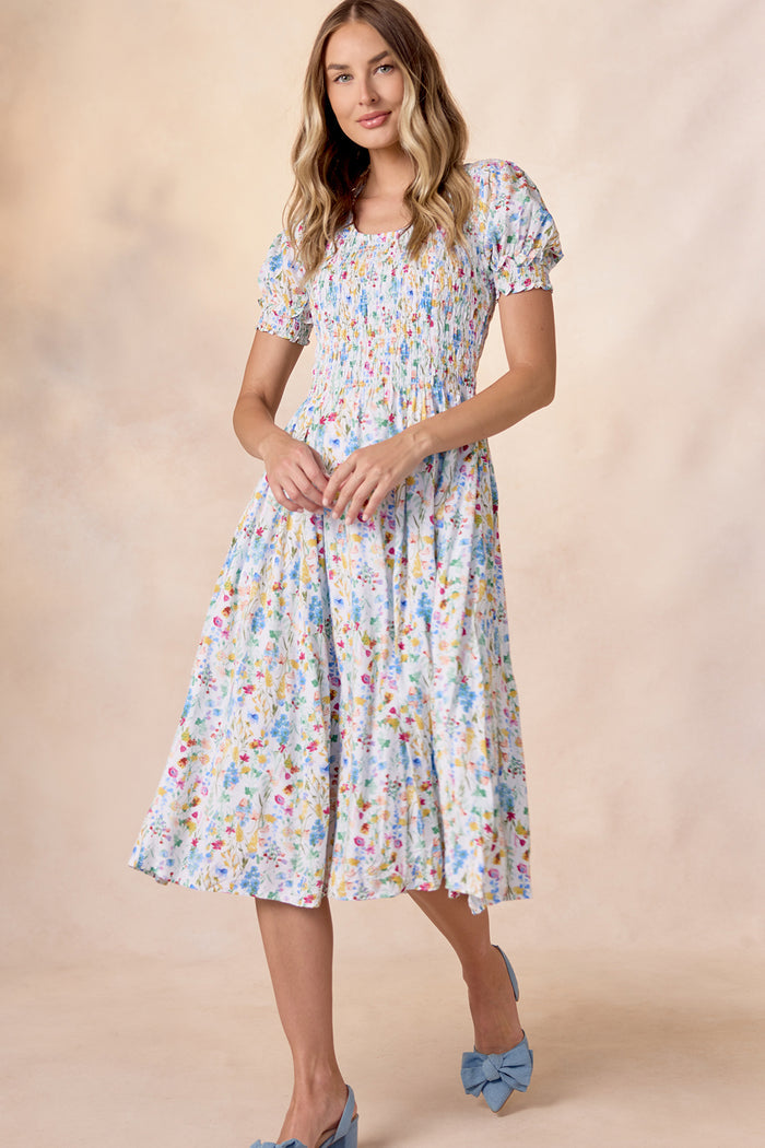 A woman wears the Ivy City Co Amelie Dress in Floral Fields—a nursing-friendly light blue floral midi with puff sleeves—paired with blue heels with bows, standing against a softly lit beige background.