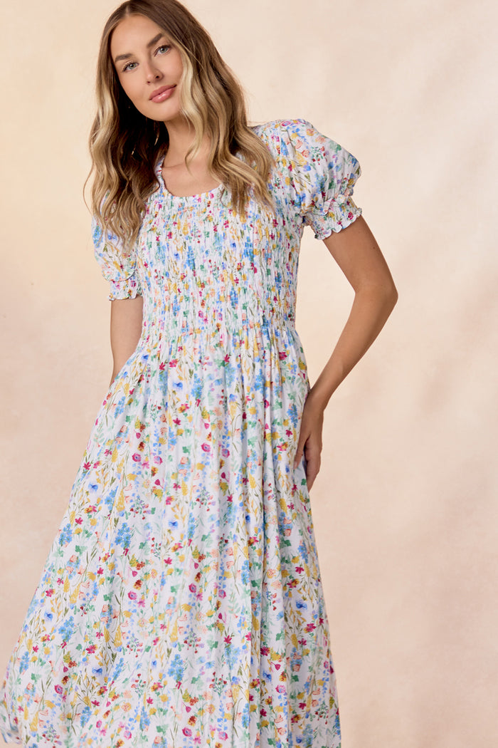 A woman in the Ivy City Co Amelie Dress in Floral Fields, a nursing-friendly light blue floral midi with smocked bodice and puff sleeves, stands against a soft beige background, smiling gently at the camera.