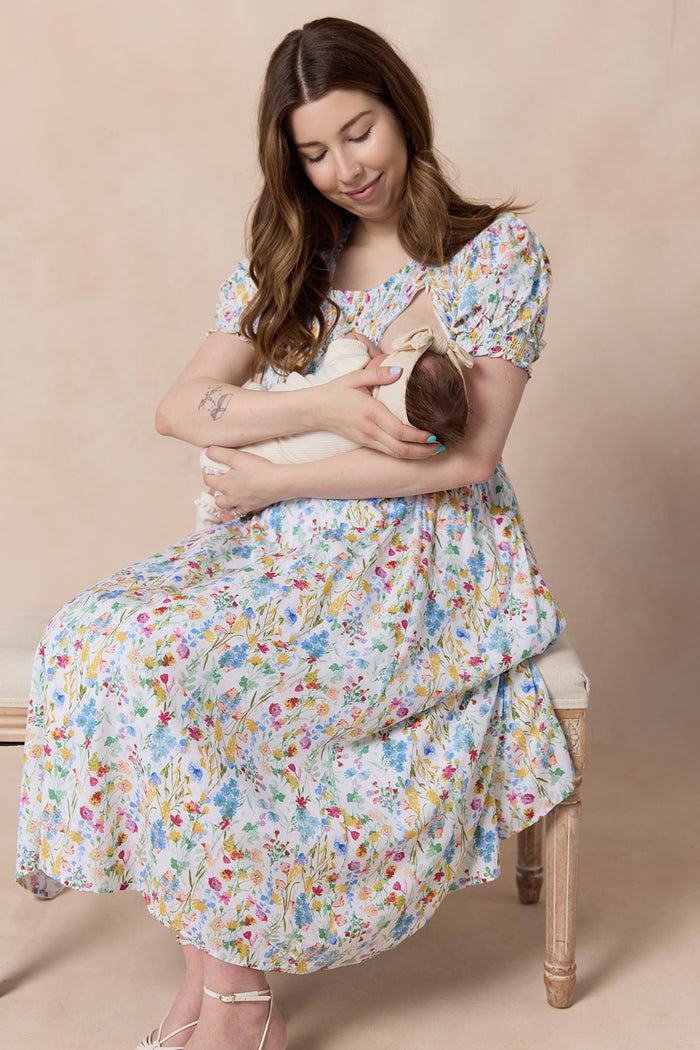 A woman wears the Ivy City Co Amelie Dress in Floral Fields, sitting on a bench and smiling at a baby wrapped in a cream blanket, set against a neutral background.