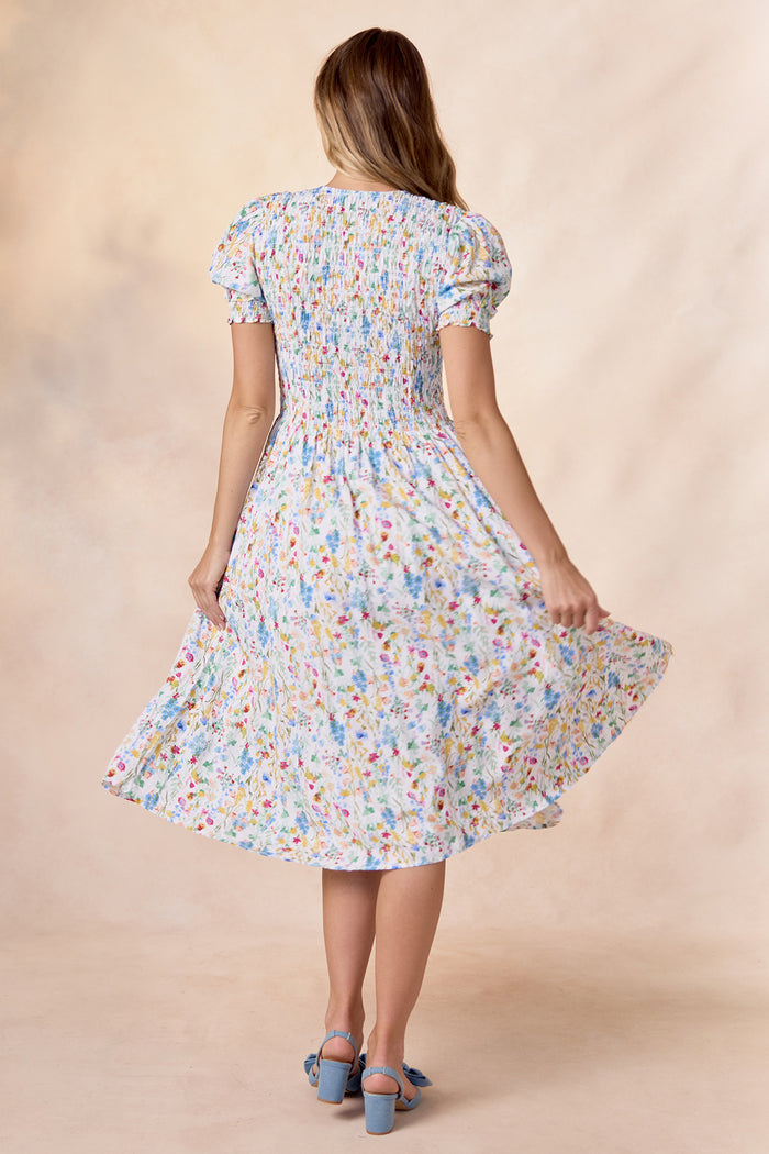 A woman with long hair faces away, wearing the Ivy City Co Amelie Dress in Floral Fields—a nursing-friendly floral midi with puff sleeves—paired with blue heeled shoes with bows. She lifts the skirt to highlight its flowy feel and vibrant print.