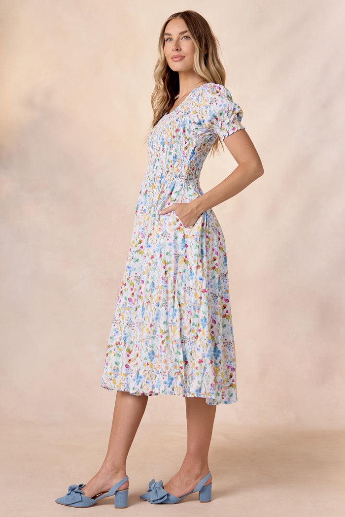 A woman stands in Ivy City Co's Amelie Dress in Floral Fields—a light blue floral midi with pockets and puff sleeves—paired with blue block-heeled bow shoes, set against a neutral beige background.