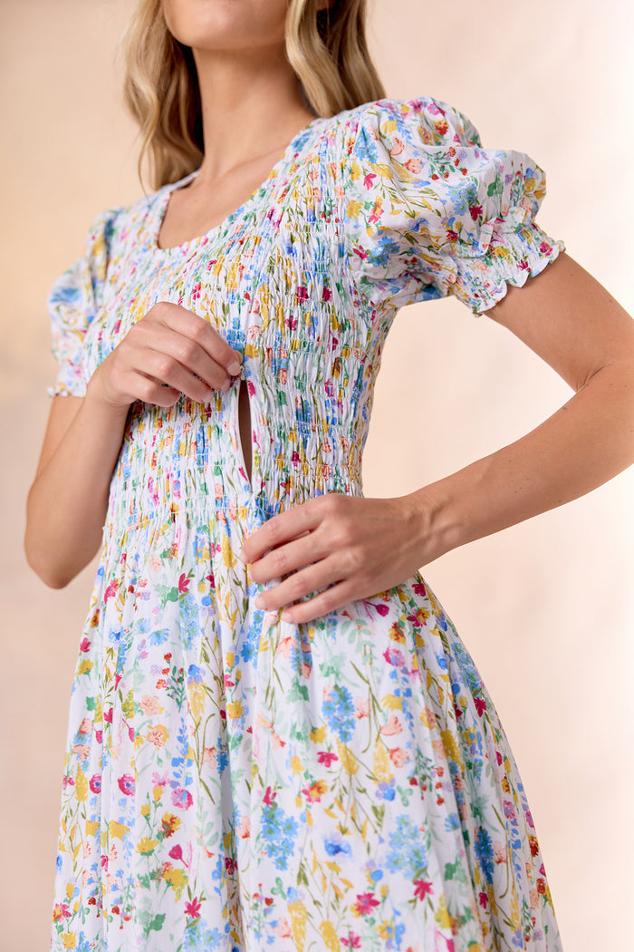 A woman wears the Ivy City Co Amelie Dress in Floral Fields, highlighting its discreet nursing-friendly access with a hidden side zipper beneath the smocked bodice and short puff sleeves.