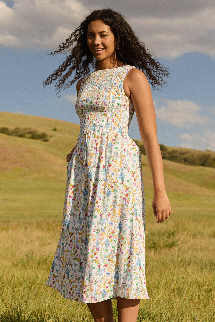 A woman stands in a grassy field wearing the Ivy City Co Ana Dress in Floral Fields—a sleeveless, floral midi with a smocked bodice. She smiles under a blue sky, accessorized with hoop earrings. Chic and maternity friendly.