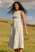 A woman stands in a grassy field wearing the Ivy City Co Ana Dress in Floral Fields—a sleeveless, floral midi with a smocked bodice. She smiles under a blue sky, accessorized with hoop earrings. Chic and maternity friendly.