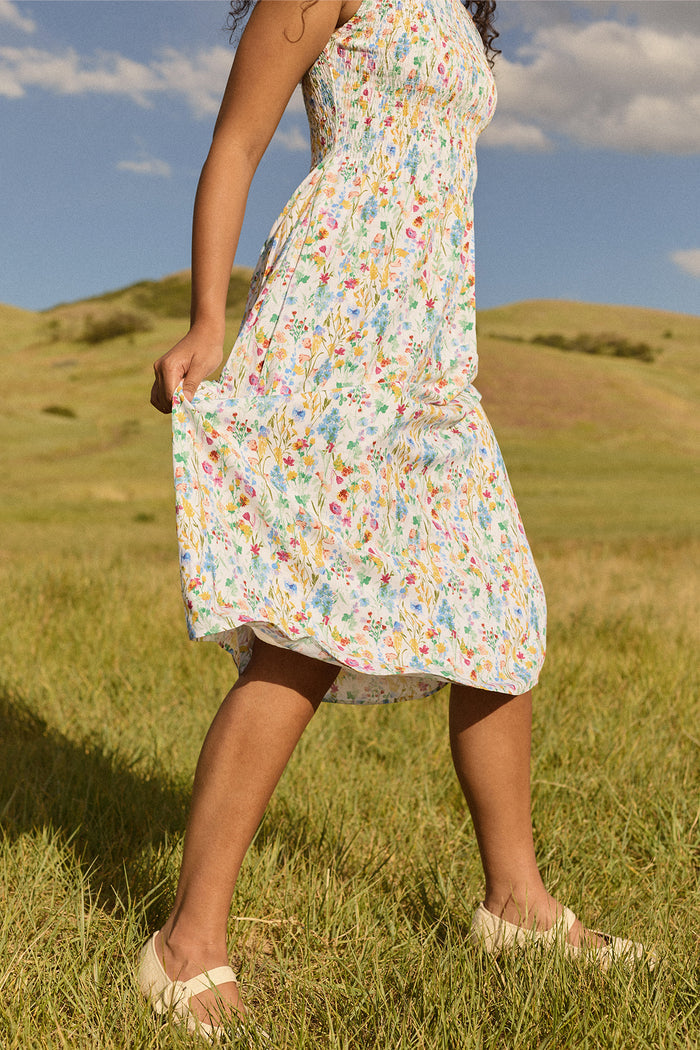 A person wearing the Ana Dress in Floral Fields by Ivy City Co walks barefoot on green grass in a sunny, open field with hills and a blue sky in the background.