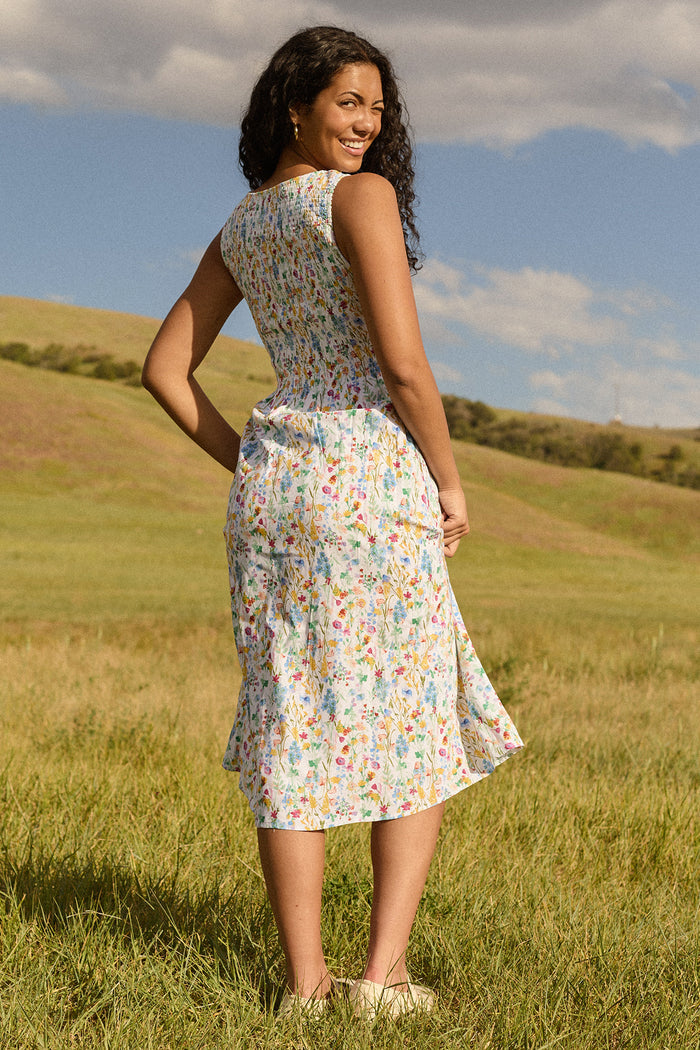 A woman with curly hair smiles over her shoulder at the camera while standing on grass, wearing the Ana Dress in Floral Fields by Ivy City Co. Green hills and a blue sky create a scenic outdoor backdrop.