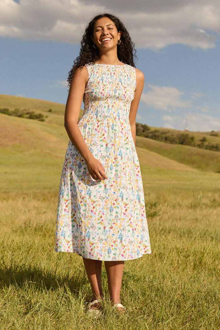 A smiling woman with curly hair stands on grass, wearing the Ana Dress in Floral Fields by Ivy City Co. Rolling green hills and a blue sky with clouds form the backdrop.