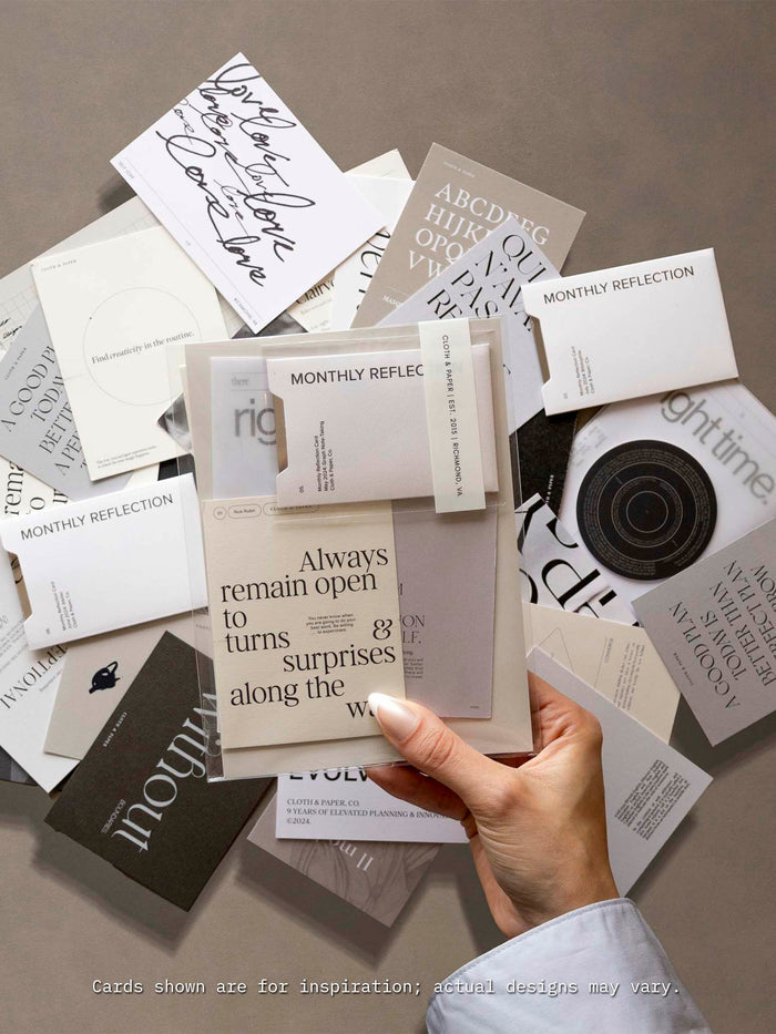 A hand holds a neutral-toned journaling card that reads ‘Always remain open to turns & surprises along the way’ above a flat lay of assorted Cloth & Paper journaling cards in minimalist fonts and designs, showcasing the variety included in the Assorted Journaling Card Set.