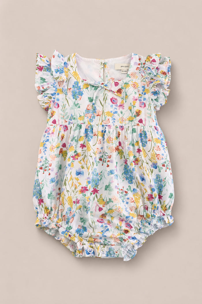 The Ivy City Co Baby Ana Romper in Floral Fields features ruffled, flutter sleeves and a charming floral print in pink, blue, yellow, and green—perfect for summer and shown on a neutral background.