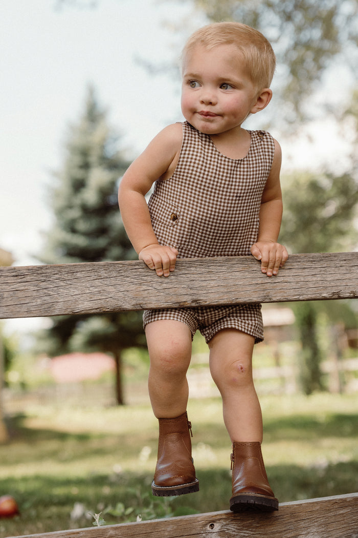 A toddler wearing the Ivy City Co Baby Boy Thomas Romper in Brown Gingham and boots sits on a wooden fence outdoors, smiling with trees and greenery in the background.