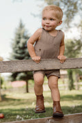 A toddler wearing the Ivy City Co Baby Boy Thomas Romper in Brown Gingham and boots sits on a wooden fence outdoors, smiling with trees and greenery in the background.