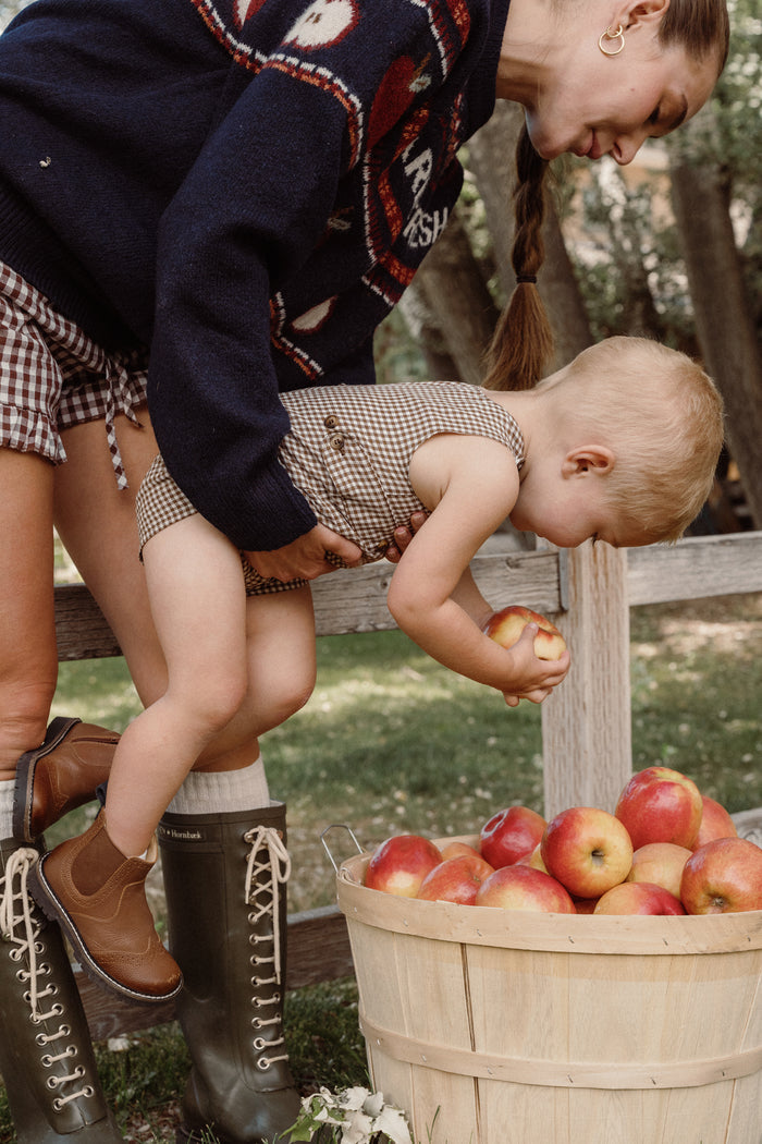 A woman helps a small child pick apples from a basket outdoors. Both wear plaid outfits; the little one is dressed in the adorable Ivy City Co Baby Boy Thomas Romper in Brown Gingham, crafted from soft woven cotton. The woman smiles and wears boots as they stand near a wooden fence, surrounded by green grass and trees.