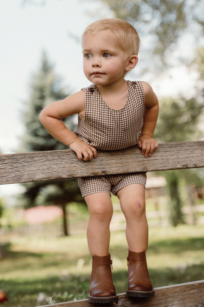 A young child dressed in the Ivy City Co Baby Boy Thomas Romper in Brown Gingham and brown boots sits on a wooden fence outdoors, gazing to the side. The woven cotton romper showcases a charming checkered gingham pattern, with trees and grass softly blurred in the background.