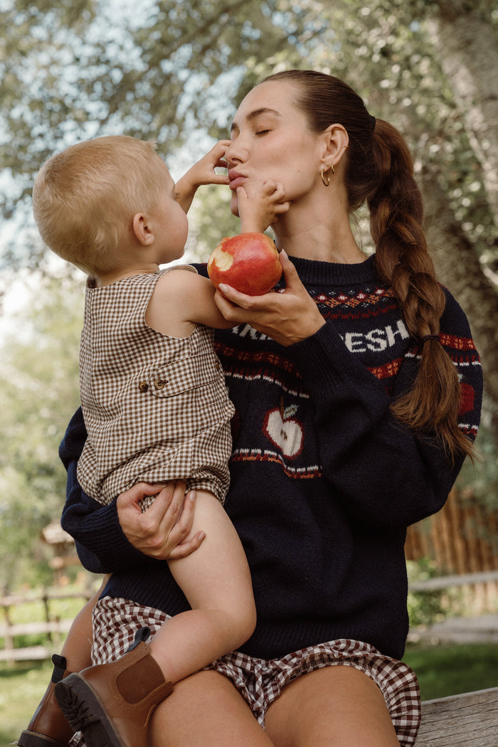 A woman sits outdoors with a young child on her lap, holding an apple. The child, dressed in the Ivy City Co Baby Boy Thomas Romper in Brown Gingham, gently touches her face while she smiles lovingly.
