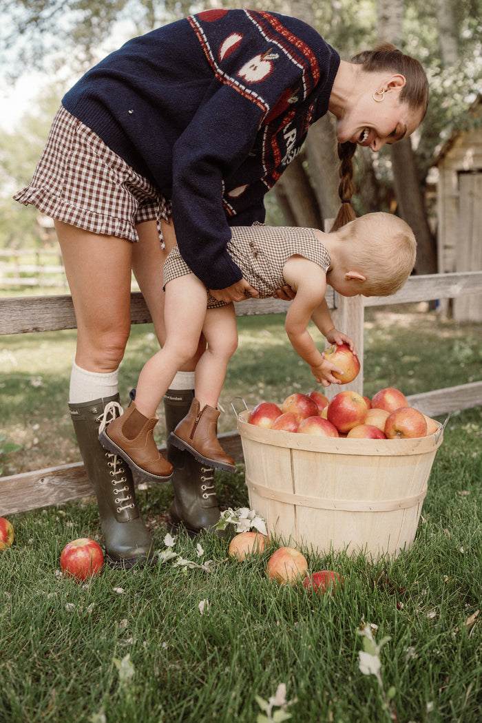A woman helps a young child, dressed in the Ivy City Co Baby Boy Thomas Romper in Brown Gingham, reach into a wooden basket full of apples outdoors. Fallen apples and leaves are scattered on the grass around them.