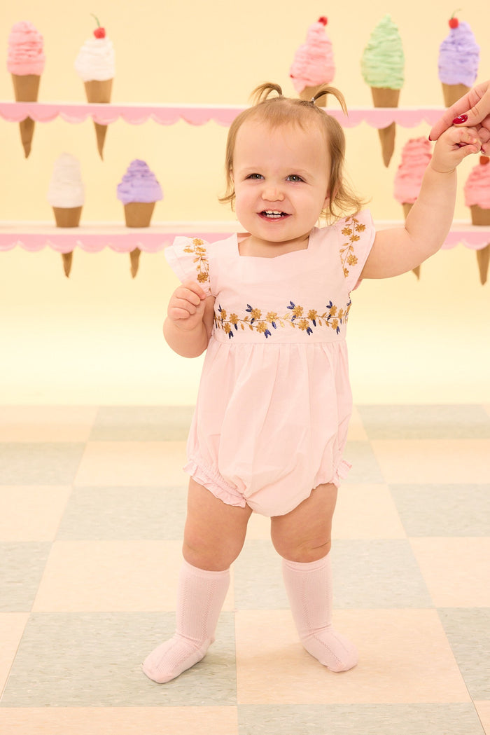 A smiling toddler wears the Ivy City Co Baby Hattie Romper in Embroidered Spring Floral with socks, holding an adult’s hand as she stands on a checkered floor decorated with colorful ice cream cone designs. The cotton romper’s delicate embroidered spring floral details add charm to her playful outfit.