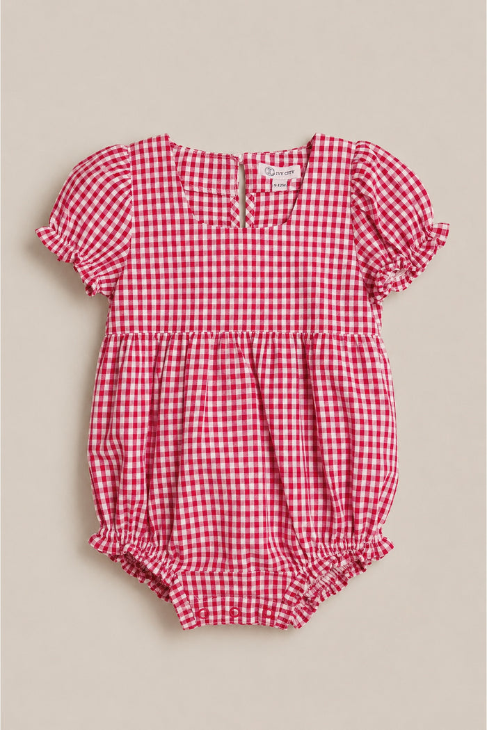 The Baby Jessie Romper in Red Gingham by Ivy City Co features short puff sleeves, ruffled edges, a gathered waist, back buttons, and elastic leg openings—ideal for the Fourth of July. Shown laid flat on a neutral background.