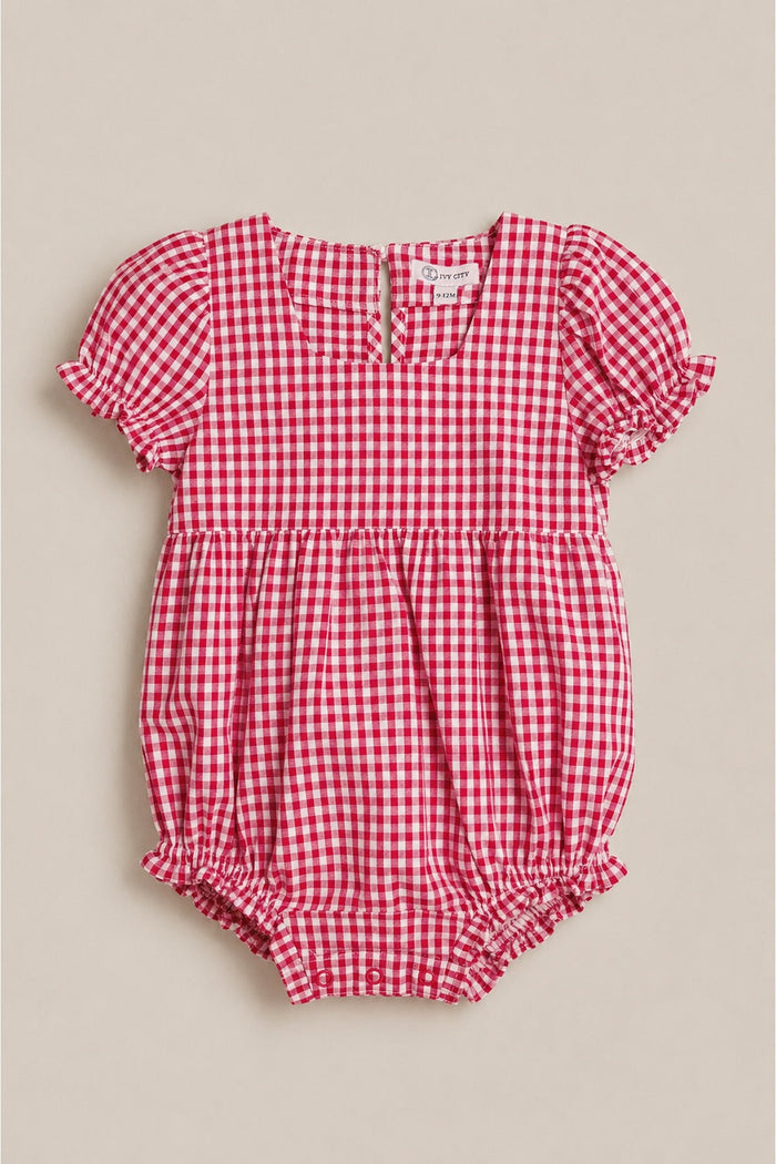 The Baby Jessie Romper in Red Gingham by Ivy City Co features a charming red and white gingham pattern, short puff sleeves, ruffle trim, and button details on the back. Displayed on a light beige background, this classic romper is perfect for an adorable baby Fourth of July outfit.