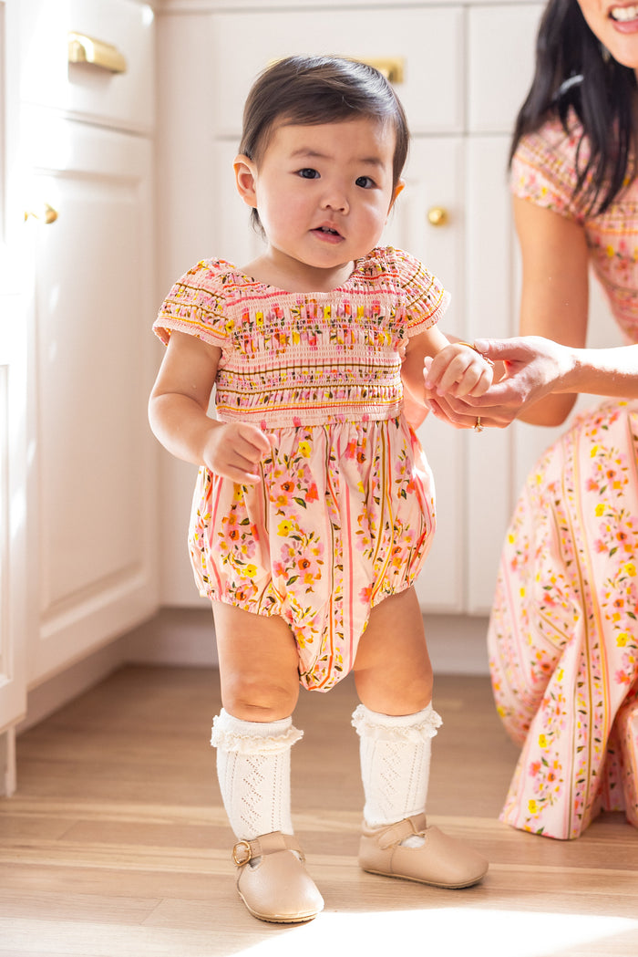 A toddler wears the Ivy City Co Baby Marlow Romper in Peach Handpainted Flora Stripe with knee-high white socks, standing on a wooden floor and holding an adult’s hand. Both sport matching mommy and me outfits in a bright, sunlit room.
