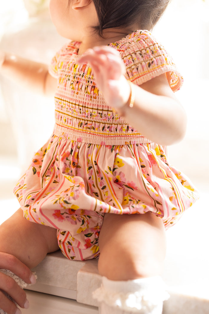 A baby in the Ivy City Co Baby Marlow Romper in Peach Handpainted Flora Stripe sits facing away, supported by an adult hand and bathed in sunlight—a sweet moment ideal for matching mommy and me outfits.