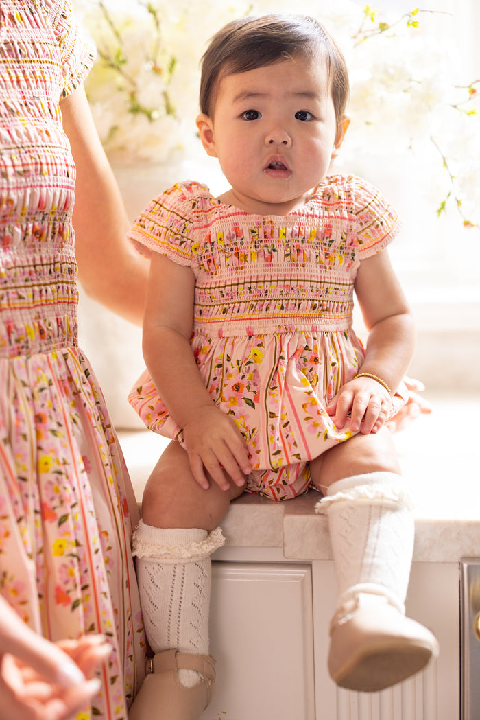 A toddler wears the Ivy City Co Baby Marlow Romper in Peach Handpainted Flora Stripe with white knee-high socks and beige shoes, sitting on a counter next to an adult in a matching outfit against a backdrop of soft, bright flowers.