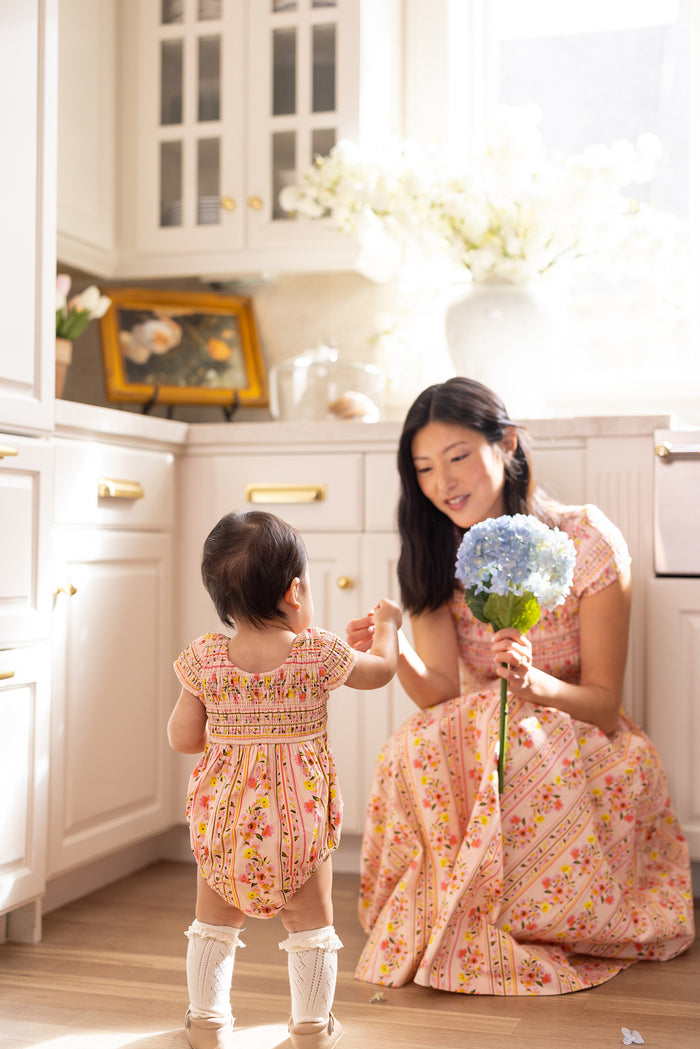 A woman kneels in a sunlit kitchen, smiling at her toddler in the Ivy City Co Baby Marlow Romper in Peach Handpainted Flora Stripe, as the child reaches for her blue hydrangea bouquet.