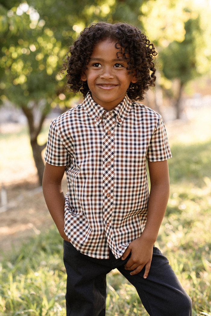A young child with curly hair smiles while standing outdoors, wearing the Ivy City Co Boys James Shirt in Chestnut Gingham—a brown and white checkered cotton shirt—paired with dark pants, with trees and sunlight in the background.