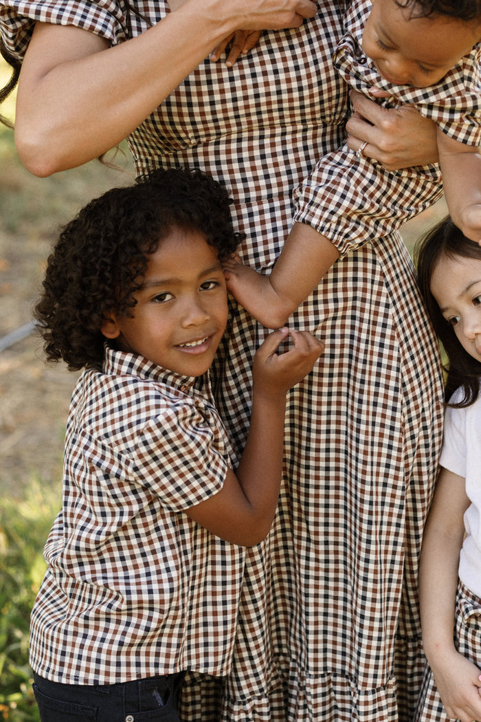 Four family members—three children and an adult—all dressed in plaid clothing from Ivy City Co, stand close together outdoors. One child hugs the adult, while another, wearing the Boys James Shirt in Chestnut Gingham, holds the adult’s arm; a third child leans in from the right, partially visible. The soft cotton shirts contribute to this cozy family moment.