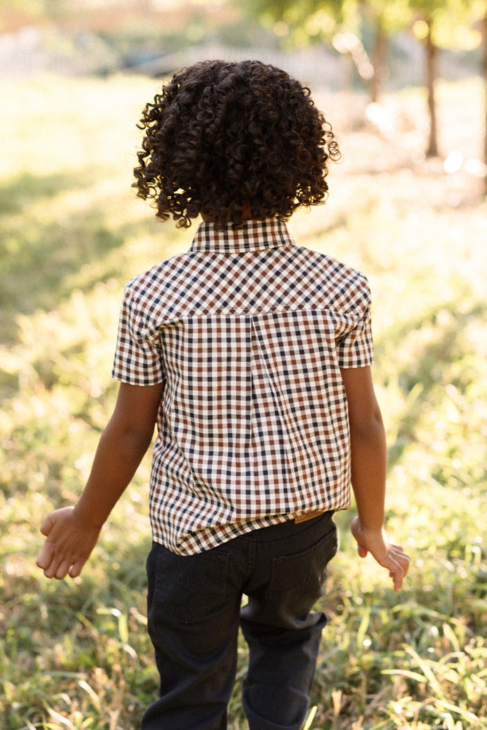 A young child with curly hair, wearing the Ivy City Co Boys James Shirt in Chestnut Gingham and dark pants, stands outdoors on a grassy area with sunlight filtering through the trees, facing away from the camera.