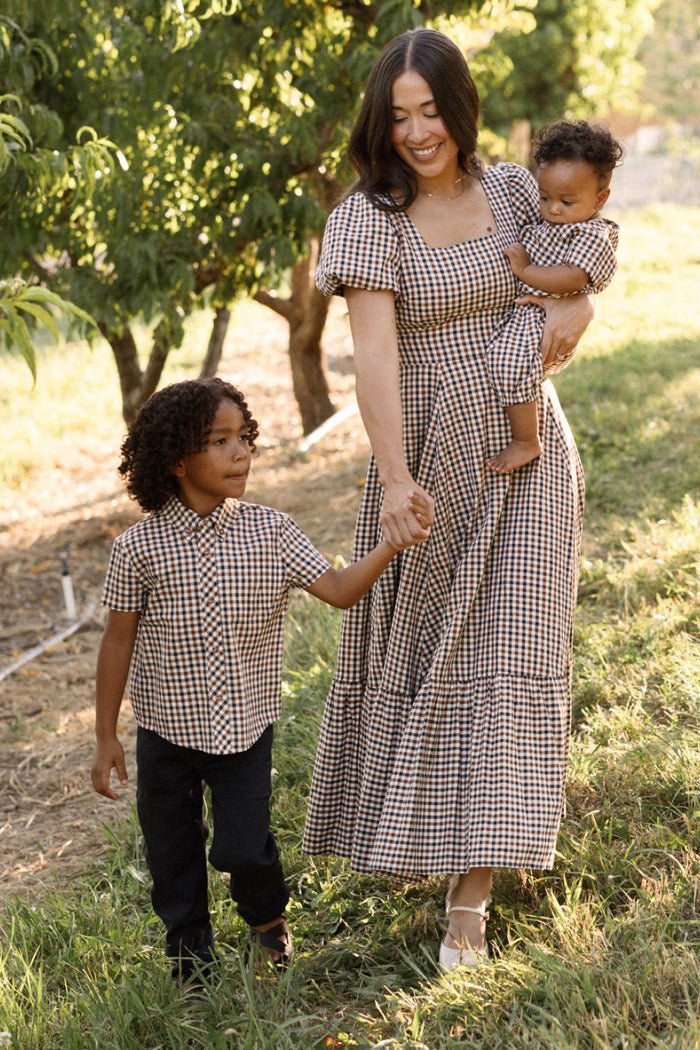 A woman and two children, all dressed in coordinated outfits featuring the Ivy City Co Boys James Shirt in Chestnut Gingham, walk together outdoors on a sunny day, surrounded by trees and grass.