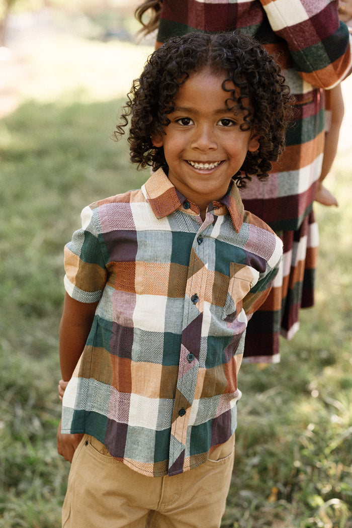 A smiling child with curly hair stands outdoors on grass, wearing the Ivy City Co Boys James Shirt in Maize Gingham with short sleeves and tan pants, while another person in a matching plaid cotton-blend outfit is partially visible behind.