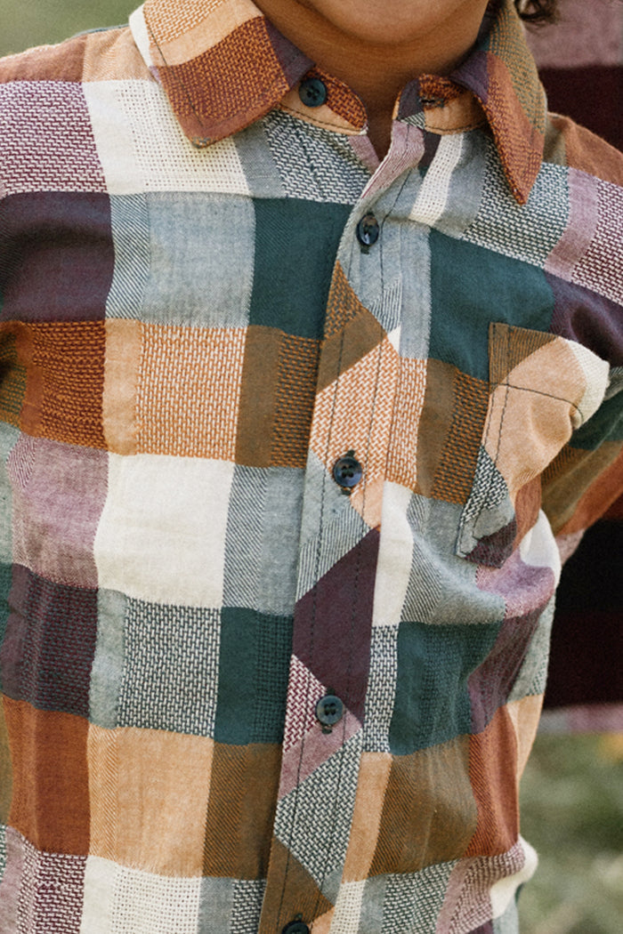 A person is seen wearing the Ivy City Co Boys James Shirt in Maize Gingham—a vibrant short-sleeve shirt featuring a colorful plaid pattern in shades of orange, green, cream, and purple. Only the torso and part of the chin are visible. Crafted from a soft cotton-blend fabric, it offers comfort and durability.