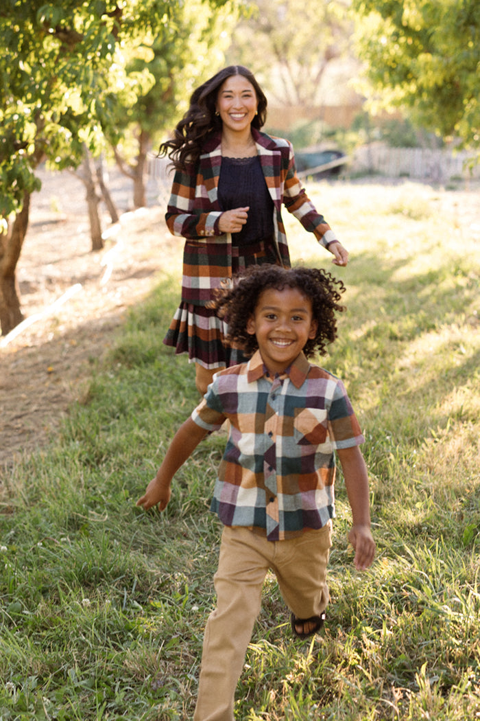 A smiling child, dressed in Ivy City Co’s Boys James Shirt in Maize Gingham—a short-sleeve style crafted from a soft cotton blend—runs ahead of a smiling woman down a grassy tree-lined path. Both wear plaid outfits in earthy tones, creating a bright and outdoorsy scene.