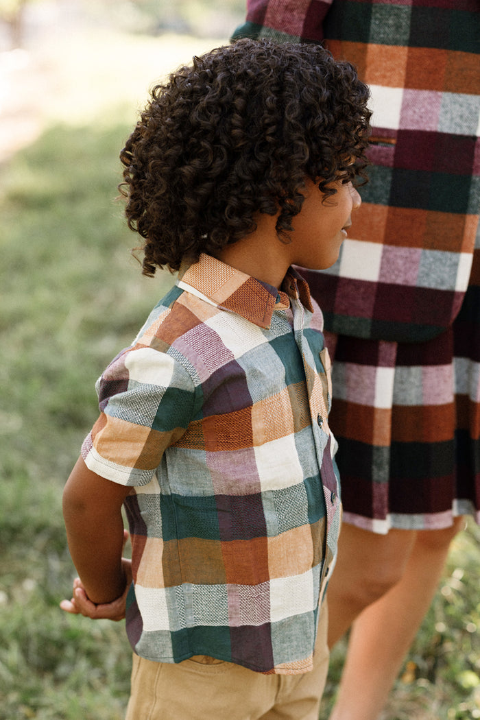 A young child with curly hair wears the Ivy City Co Boys James Shirt in Maize Gingham, featuring short sleeves and crafted from a comfortable cotton-blend fabric, paired with tan shorts while standing outdoors beside an adult in a matching gingham outfit.