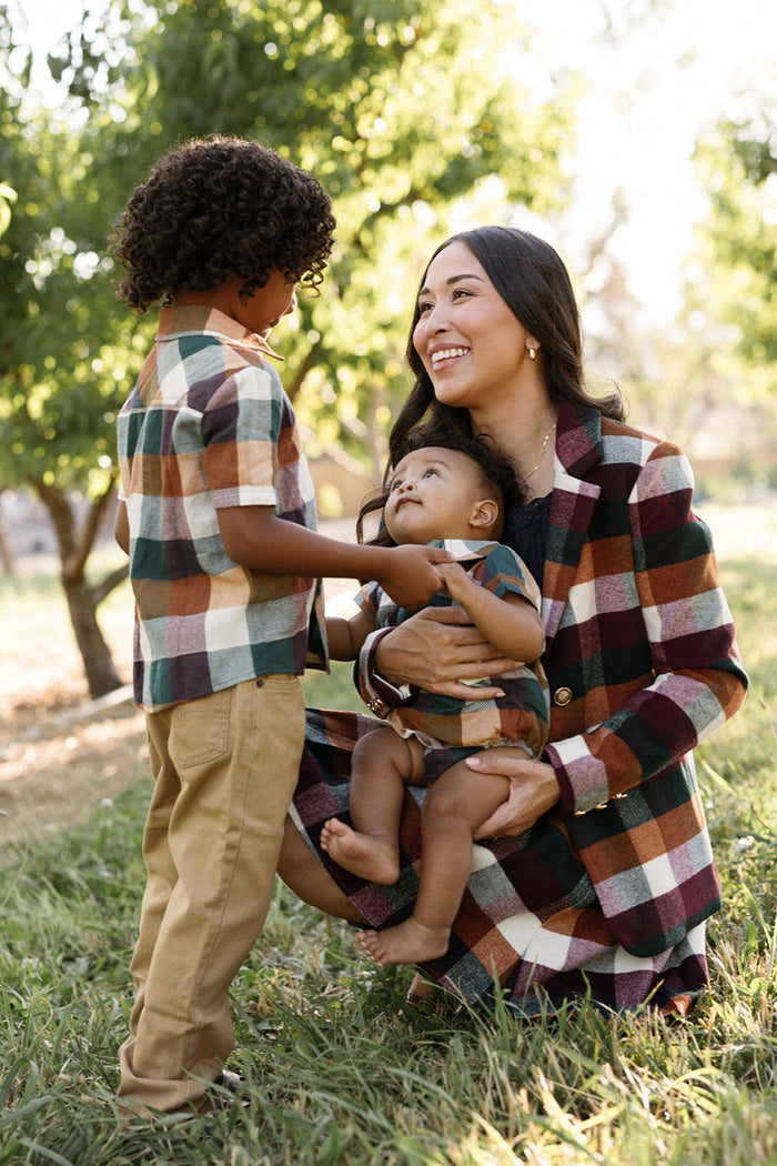 A woman kneels on the grass, smiling at a young boy wearing the Ivy City Co Boys James Shirt in Maize Gingham with short sleeves and holding a baby. All three are dressed in matching plaid outfits made from soft cotton-blend fabric, with trees in the background.