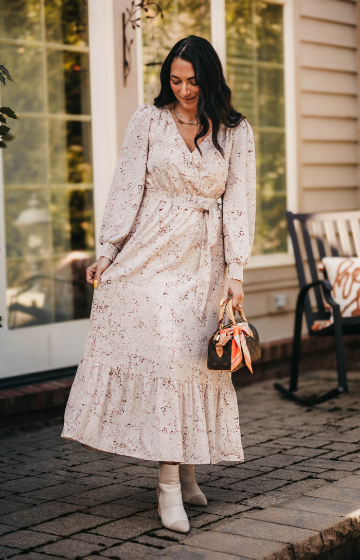 A woman wearing a long-sleeve, belted midi dress with a delicate floral print in beige and pink, standing on a brick patio in front of a house. She is holding a small brown handbag with a pink ribbon and wearing white ankle boots.