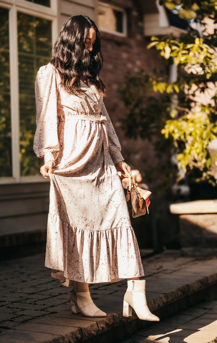A woman wearing a light beige floral midi dress with long sleeves and a tiered ruffle hem, paired with white heeled ankle boots and holding a small tan handbag, standing outdoors on a stone walkway.