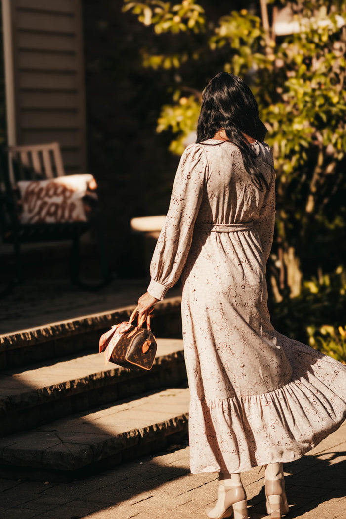 A woman viewed from behind wearing a long-sleeve beige maxi dress with a small floral print, walking down stone steps outdoors while holding a small handbag.