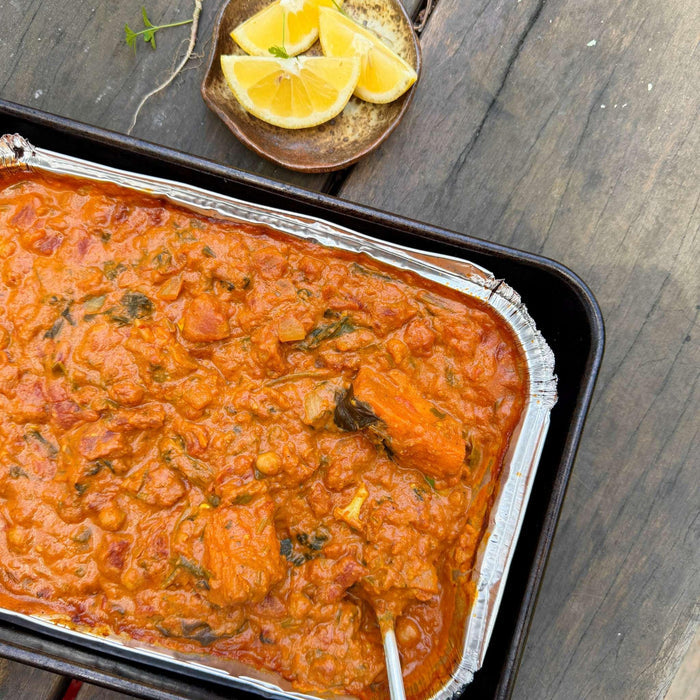 Organic ready meal with rich tomato sauce, chickpeas, and greens in foil tray, served with lemon wedges on rustic wood.